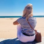 Blondynka w Maroku siedzi odwrócona plecami. Przed sobą ma ocean i plażę. Nosi koszulkę w marynarskie paski i białe spodnie.
