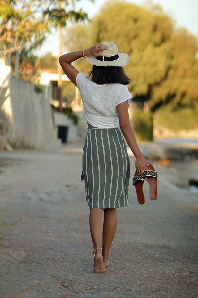 Kobieta ubrana w białą bluzkę i spódnicę w paski spaceruje, trzymając w rękach klapki.